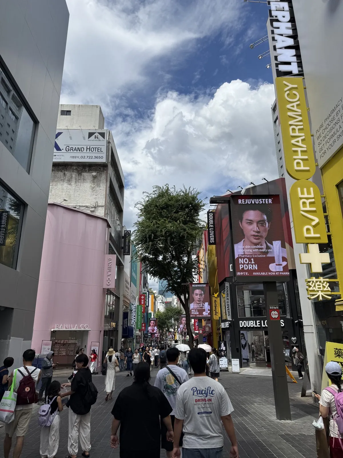A crowded Myeongdong shopping street with a large yellow pharmacy sign, a skincare advertisement on a screen beside it, cosmetics shops along both sides and a hotel sign above the rooftops