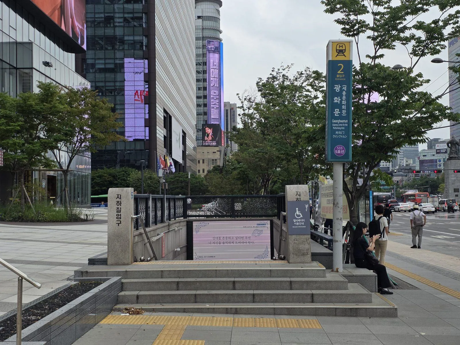 A Seoul subway entrance at street level, stairs leading down beside a tall sign giving the exit number, with a main road and shopfronts behind