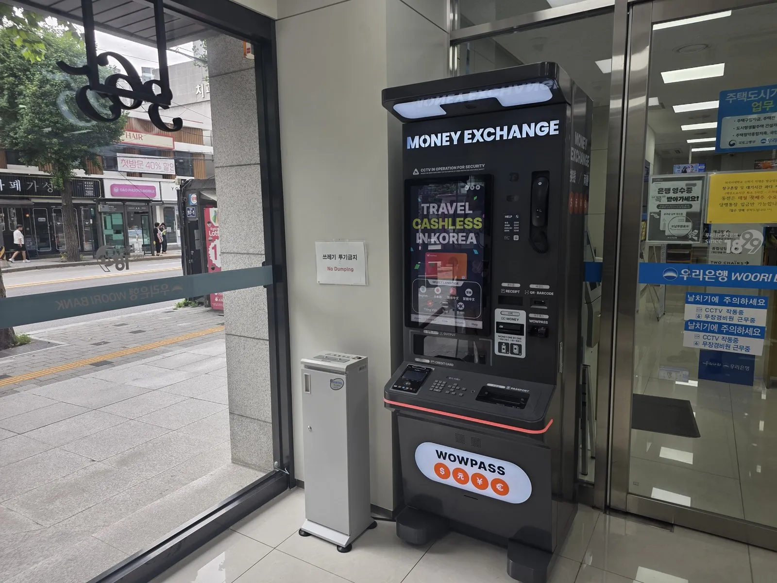 A WOWPASS money exchange kiosk inside a bank entrance, MONEY EXCHANGE lit above the screen and a WOWPASS panel below it